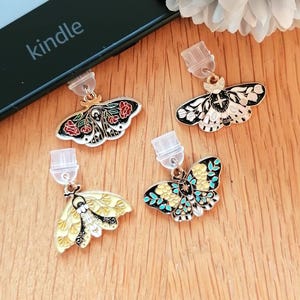 May include: Four pairs of moth-shaped earrings with colourful enamel designs. The earrings are in gold and silver tones, with red, black, yellow, and blue accents. The word "kindle" is visible on a black electronic device.