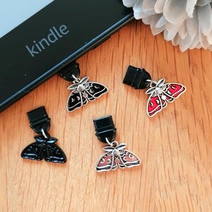 Puede incluir: Cuatro colgantes decorativos con forma de polilla en negro, rojo y marrón, con detalles plateados. Cada colgante tiene un clip negro. Los colgantes se muestran sobre una superficie de madera cerca de un lector Kindle.