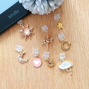 Può includere: Una collezione di piccoli ciondoli decorativi con motivi celesti e a forma di cuore. I ciondoli includono un sole, stelle, luna e nuvole, in oro, rosa e bianco. Un lettore Kindle e un fiore azzurro sono anche nell'immagine.