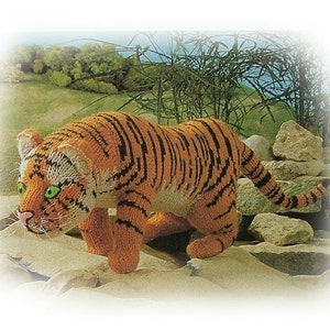 Peut inclure: Figurine de tigre tricoté avec des rayures orange et noires, des yeux verts et un nez rose. Le tigre fait à la main est posé sur une surface sablonneuse avec un fond de rochers et de verdure. C'est une sculpture souple.