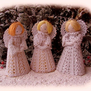 May include: Three handmade angel figurines with white crocheted dresses and wings, set in a snowy scene. Each angel has yarn hair in different colours: orange, yellow, and blonde. The angels are decorated with gold and silver accents, ideal for festive decorations.