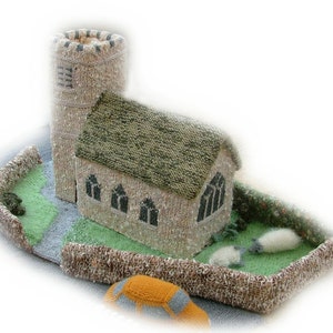 Könnte beinhalten: Eine handgefertigte Miniatur-Kirchszene mit einem Steinturm, einem Kirchengebäude und einer grünen Wiese mit zwei Schafen. Die Szene ist von einer gestrickten Steinmauer umgeben. Das Dach der Kirche ist grün.