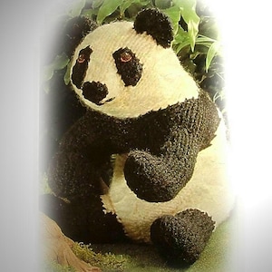 May include: A handmade panda bear plush toy with black and white knitted details. The panda has black ears, eyes, and limbs, with a white face and body. The plush is set against a backdrop of green foliage.