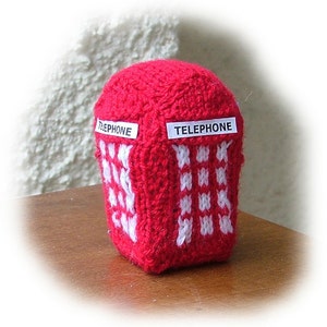 May include: A knitted red telephone booth with white accents. The word "TELEPHONE" is displayed on two white rectangular labels. The booth has a square shape with a rounded top and a pattern of white squares.