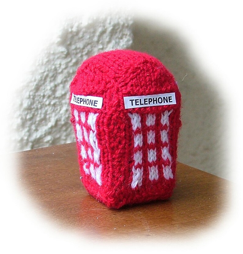 May include: A knitted red telephone booth with white accents. The word "TELEPHONE" is displayed on two white rectangular labels. The booth has a square shape with a rounded top and a pattern of white squares.