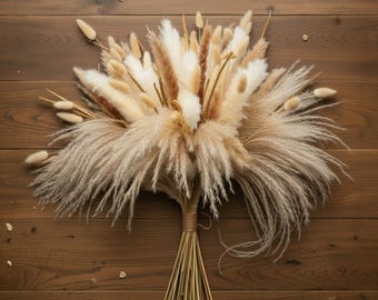 Può includere: Un bouquet di fiori secchi con erba della pampa, code di coniglio e altri elementi secchi nei toni del beige, crema e marrone. Gli steli sono legati con spago, creando un'estetica naturale. Il bouquet è disposto su una superficie di legno.