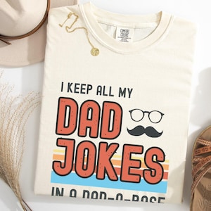 May include: A cream-colored t-shirt featuring the text "I KEEP ALL MY DAD JOKES IN A DAD-A-BASE" in black and red lettering. The design includes a graphic of black eyeglasses and a mustache, set against a background of horizontal stripes.