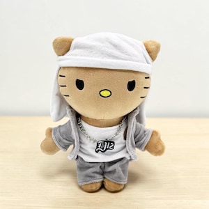 May include: A plush cat toy dressed in a white beanie, grey jacket, and trousers. The cat has a silver chain necklace with a "D12" pendant. The cat's face features black eyes, whiskers, and a yellow nose.