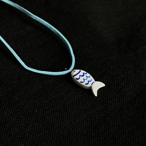 Könnte beinhalten: Halskette mit hellblauem Band und einem Anhänger in Form eines weiß-blauen Fisches. Der Fisch hat blaue wellenförmige Muster und einen weißen Schwanz. Die Kette liegt auf schwarzem Hintergrund.