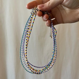 May include: A multi-strand necklace featuring small, vibrant, multi-colored glass seed beads in shades of blue, yellow, pink, purple, and white. The necklace includes a metallic silver-toned clasp and an adjustable chain extension for length.