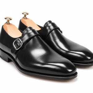 Könnte beinhalten: Ein Paar schwarze Lederschuhe mit Monk-Strap-Riemen und silbernen Schnallen. Die Schuhe werden auf hölzernen Schuhspannern vor weißem Hintergrund präsentiert. Das Leder hat ein glänzendes Finish.
