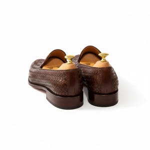 May include: A pair of dark brown woven leather loafers featuring a textured basketweave pattern, a low stacked heel, and light-coloured wooden shoe trees with polished gold-tone metal knobs inserted into the back of each shoe.