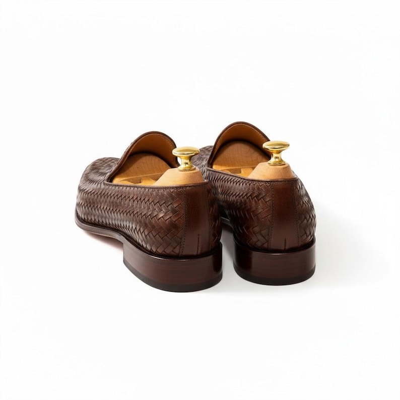 May include: A pair of dark brown woven leather loafers featuring a textured basketweave pattern, a low stacked heel, and light-coloured wooden shoe trees with polished gold-tone metal knobs inserted into the back of each shoe.