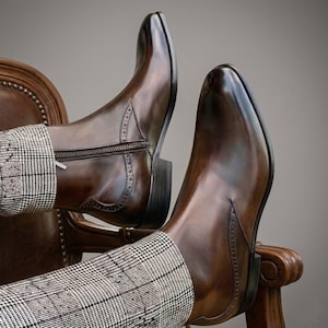 Puede incluir: Un par de botas de cuero marr&oacute;n pulido con punta cuadrada y cremallera lateral. Las botas tienen costuras decorativas y se usan con pantalones estampados. Las botas descansan sobre una silla de madera.