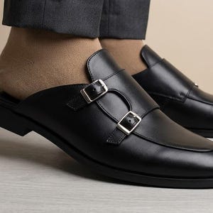 May include: A pair of black leather mule loafers featuring a double monk strap design with silver-toned metal buckles. The footwear has a smooth finish, a rounded toe, and an open back, worn with beige socks and dark grey trousers.