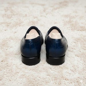 May include: A pair of navy blue leather loafers with a crocodile texture. The shoes have a low heel and are displayed on a white, fluffy rug. Shoe trees are inserted inside the shoes.