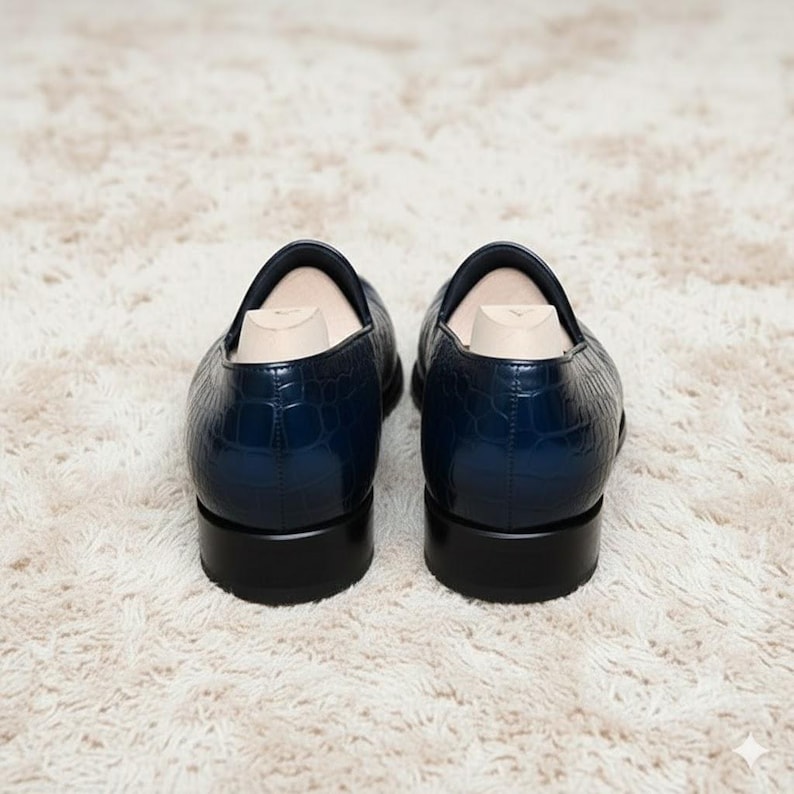 May include: A pair of navy blue leather loafers with a crocodile texture. The shoes have a low heel and are displayed on a white, fluffy rug. Shoe trees are inserted inside the shoes.