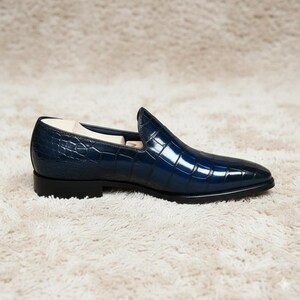 May include: A single, navy blue leather loafer with a crocodile texture. The shoe has a pointed toe and a low heel. The shoe is displayed on a cream-coloured, textured surface.