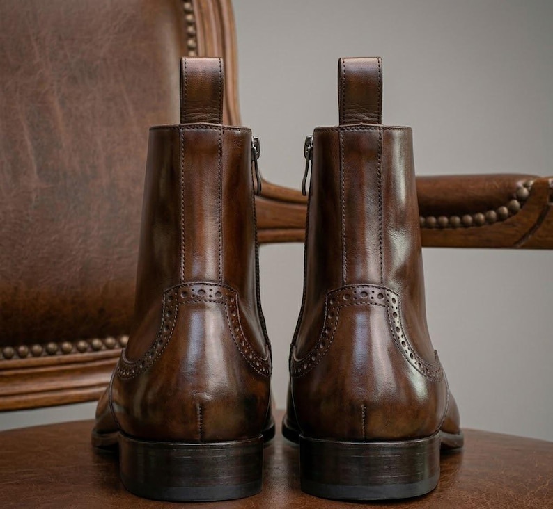 Puede incluir: Dos botines de cuero marr&oacute;n con tiradores y cremalleras laterales. Los botines tienen perforaciones decorativas y est&aacute;n sobre un fondo borroso de una silla de cuero.