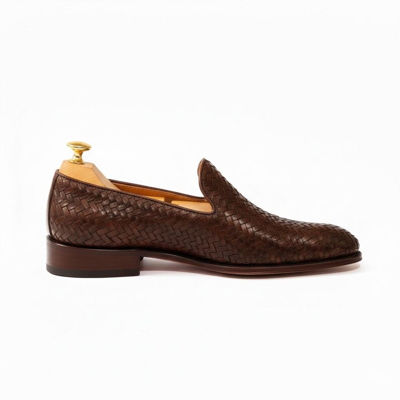 May include: A dark brown woven leather loafer with a low heel and a smooth dark brown sole. The shoe features a detailed basket-weave pattern across the entire upper surface and includes a light-coloured wooden shoe tree with a gold-toned metal knob.