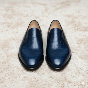 May include: A pair of navy blue leather loafers with a textured, crocodile-like pattern. The shoes have a classic slip-on design with a rounded toe and a black sole. The loafers are displayed on a cream-coloured, textured surface.