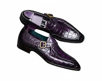 May include: A pair of purple leather dress shoes with a crocodile skin texture. The shoes have a buckle and tassel detail. The shoes are set against a white background.