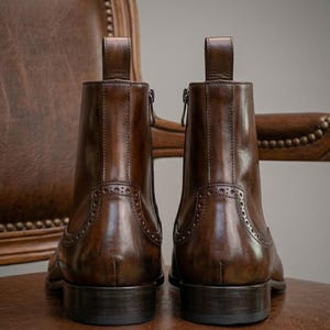 Puede incluir: Dos botines de cuero marr&oacute;n con tiradores y cremalleras laterales. Los botines tienen perforaciones decorativas y est&aacute;n sobre un fondo borroso de una silla de cuero.