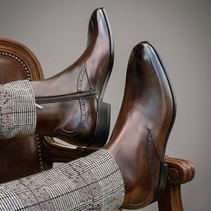 Puede incluir: Un par de botas de cuero marr&oacute;n pulido con punta cuadrada y cremallera lateral. Las botas tienen costuras decorativas y se usan con pantalones estampados. Las botas descansan sobre una silla de madera.