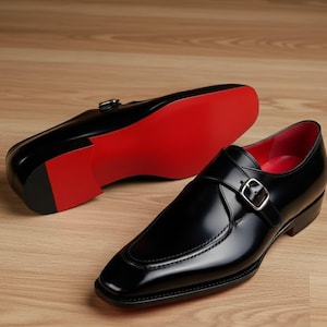 May include: Pair of black leather dress shoes with a glossy finish. The shoes feature a buckle closure and a red sole. The interior lining is also red. The shoes are displayed on a wooden surface.