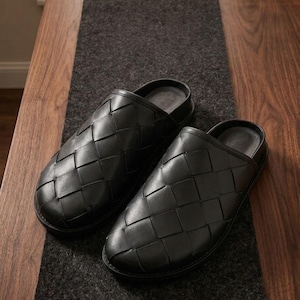 Könnte beinhalten: Ein Paar schwarze, gewebte Lederslipper. Die Slipper liegen auf einem dunkelgrauen Läufer auf einem Holztisch. Die Slipper haben ein Schlupfdesign und eine abgerundete Spitze.