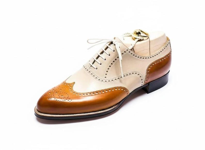 May include: A two-tone leather oxford shoe with a cream-colored upper and a brown toe and heel. The shoe features decorative perforations and white laces. The sole is black, and the shoe is on a white background.