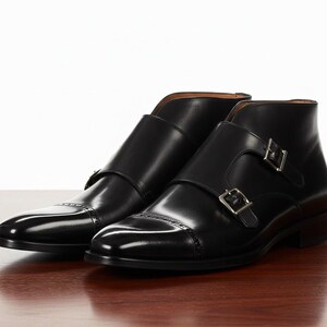 May include: A pair of black leather ankle boots with double buckle fastenings. The boots have a polished finish and a classic design, suitable for formal or business attire. The boots are displayed on a wooden surface.