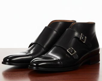 Puede incluir: Un par de botines de cuero negro con cierres de doble hebilla. Los botines tienen un acabado pulido y un diseño clásico, adecuado para vestimenta formal o de negocios. Los botines se exhiben sobre una superficie de madera.