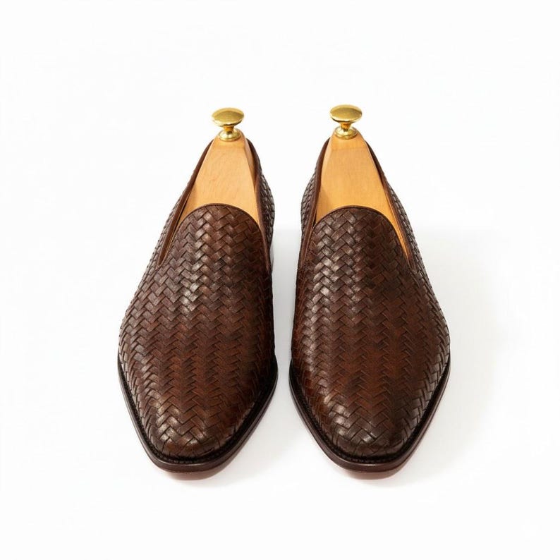 May include: A pair of dark brown woven leather loafers featuring a slip-on design with a pointed toe. Each shoe includes a light-coloured wooden shoe tree with a gold-toned metal knob handle inserted into the opening to maintain the shape.