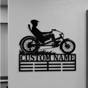 May include: Black metal recumbent bike medal display with the text "CUSTOM NAME" and horizontal bars for hanging medals. The display features a silhouette of a person riding a recumbent bike. A framed display of medals and certificates is to the right.