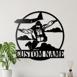 May include: Black metal wall art depicting a skydiver in mid-air, with a plane above and clouds below. The words "CUSTOM NAME" are displayed at the bottom. The art is circular and suitable for indoor display.