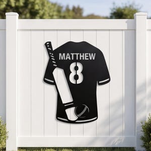 Könnte beinhalten: Schwarzes Metallschild in Form eines Cricket-Trikots mit dem Namen "MATTHEW" und der Nummer "8". Ein Cricket-Schläger und ein Ball sind links vom Trikot positioniert. Das Schild ist an einem weißen Zaun befestigt.