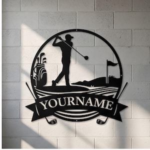 Puede incluir: Arte de pared de metal negro con temática de golf. Un golfista se representa en pleno swing dentro de un marco circular, con una bolsa de golf y una bandera visibles. La pancarta de abajo dice "YOURNAME". Dos palos de golf a cada lado.