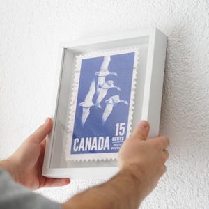 Könnte beinhalten: Ein gerahmter kanadischer Briefmarken-Klassiker. Die Briefmarke ist blau mit weißen Akzenten und zeigt ein Design von fliegenden Vögeln. Der Text "CANADA" und "15 CENTS" sind sichtbar. Der Rahmen ist weiß.
