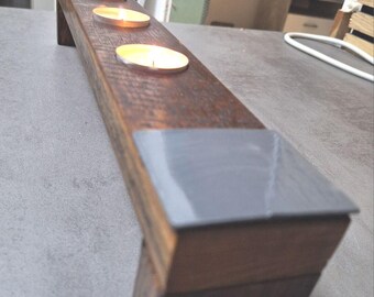 May include: A rectangular wooden candle holder with three lit tea light candles. The dark brown wood has a textured appearance, with gray metal accents on the ends. The candles are in silver metal holders.