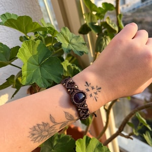 May include: A brown macrame bracelet featuring a dark purple gemstone and gold-coloured beads. The bracelet is worn on a wrist with tattoos of a sunflower and a starburst design. The background includes green leaves and sunlight.