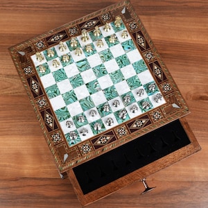 May include: A decorative chess set with a wooden frame and a drawer. The chessboard features a green and white checkered pattern with gold and silver chess pieces. The set is displayed on a wooden surface.