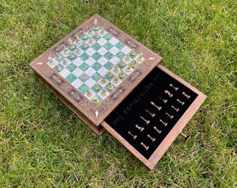 Op de afbeelding: Een houten schaakspel met een lade met schaakstukken. Het schaakbord heeft een groen-wit geblokt patroon met gouden en zilveren schaakstukken. De set ligt op een bed van groen gras.