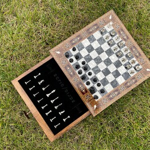 Pode incluir: Um jogo de xadrez de madeira com uma borda decorativa e uma superfície de jogo quadriculada em preto e branco. O conjunto inclui peças de xadrez pretas e prateadas e uma gaveta para armazenamento. O conjunto é exibido em uma superfície gramada.