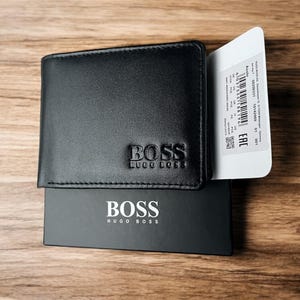 May include: Black leather wallet with the embossed BOSS Hugo Boss logo. The wallet is on top of a black box with the BOSS logo in white. A white tag with text is visible.