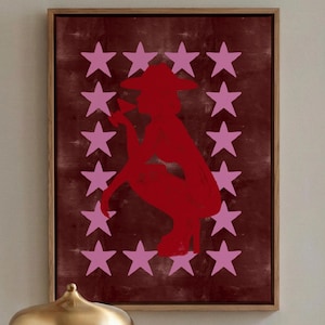 May include: A framed artwork features a red silhouette of a person in a hat, surrounded by pink stars on a burgundy background. A gold-toned lamp and a bar cart with glassware and bottles are in the foreground, creating a stylish interior scene.