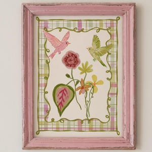 May include: A framed print featuring a pink bird and a green bird flying above a cluster of flowers and leaves. The artwork is enclosed in a distressed pink wooden frame and set against a background with a green and pink plaid border pattern.