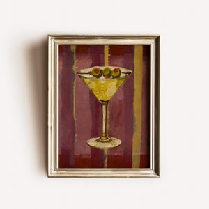 Op de afbeelding: Een ingelijst schilderij van een martini-cocktail met drie olijven. Het martini-glas is geel en de achtergrond heeft verticale strepen in bordeauxrood, goud en bruin. De lijst is goudkleurig.