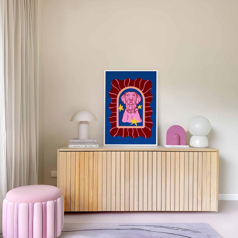 May include: A framed art print featuring a pink dog illustration on a blue background, framed by a red decorative border. Two yellow stars are included in the design. The artwork is displayed on a wooden cabinet, with a pink ottoman and decorative objects.