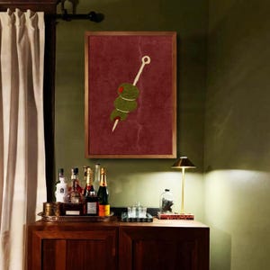 Puede incluir: Una impresión artística enmarcada que presenta una representación estilizada de tres aceitunas verdes en un palillo sobre un fondo rojo oscuro. La obra de arte está en un marco de madera y cuelga de una pared verde, lo que sugiere un bar o un comedor.